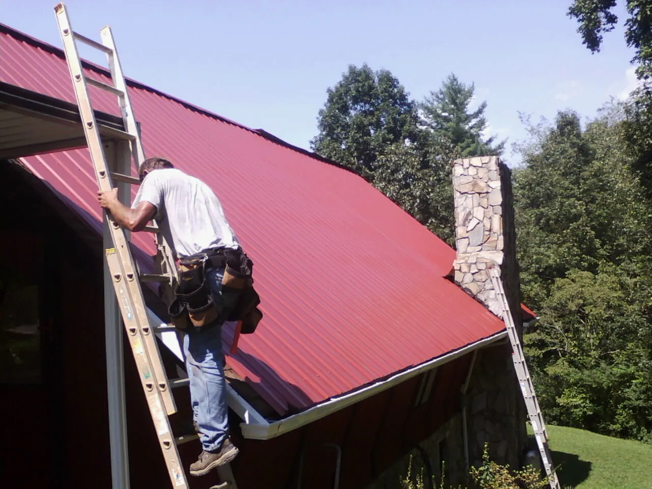 Roofer on a ladder beside a red metal roof and stone chimney — Soffit & Fascia Repair by Roofing Brothers in Hancock